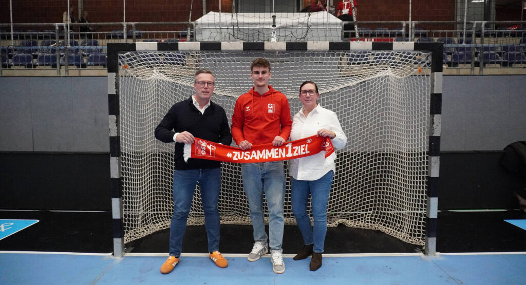 Das Bild zeigt eine Frau und einen Mann die im Auftrag von der Firma ERICH BENDER GmbH & Co. KG, da sind. In der Mitte steht ein Spieler der HSG, Sie halten gemeinsam einen Fan-schal hoch wo draufsteht "Zusammen1Ziel".